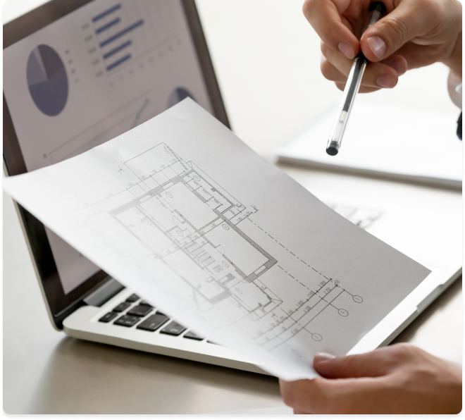 Hands working on a laptop with a blueprint and pen, indicating architectural planning.